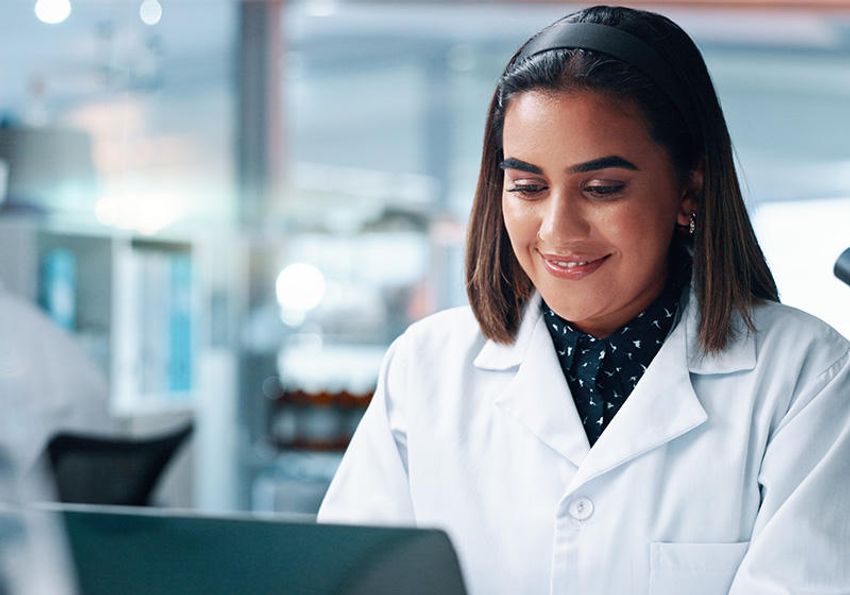 A scientist wearing a lab coat looks at her laptop while in the research laboratory