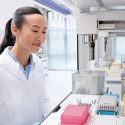 ] Image of a smiling scientist in a lab coat gazing at pipette tips and an automated pipette on a workbench.