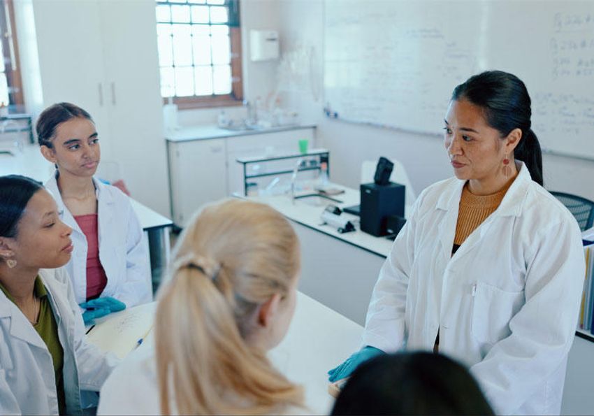 A science teacher standing at a lab bench surrounded by students in white lab coats