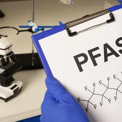 A researcher holding a clipboard with a sheet of paper that reads PFAS and includes a PFAS chemical structure
