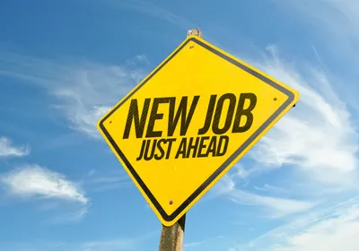Photo of a bright yellow, diamond-shaped road sign with the words “New Job Just Ahead” printed in black. 