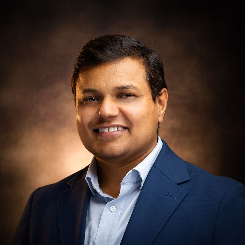 Photograph of Vishnu Tripathi, a senior scientist at Waters Biosciences, with a brown background. Tripathi has dark hair and is wearing a light blue collared shirt and dark blue dress jacket while smiling at the camera.