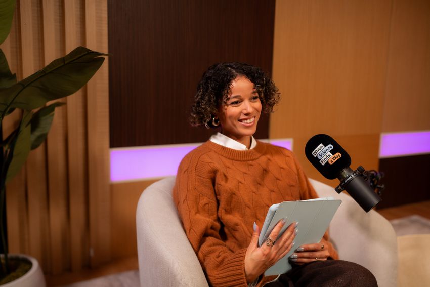 Photograph of Alisha Wainwright interviewing someone off camera and smiling. She has dark brown curly hair and is wearing an orange sweater. She’s holding a tablet and a podcasting microphone sits in front of her.