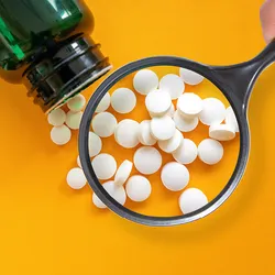 Circular white pills are shown spilling out of a green medicine bottle on a bright yellow-orange background. A magnifying glass is placed over the pills, indicating drug regulators’ inspection process for new drugs.