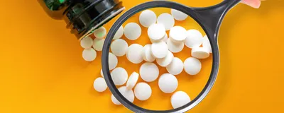 Circular white pills are shown spilling out of a green medicine bottle on a bright yellow-orange background. A magnifying glass is placed over the pills, indicating drug regulators’ inspection process for new drugs.