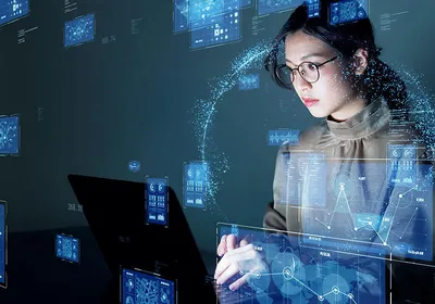 A scientist wearing glasses types on her laptop while images of graphs and other data surround her