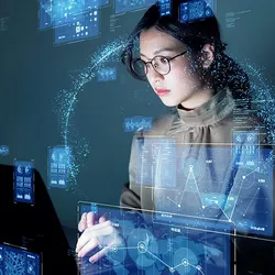 A scientist wearing glasses types on her laptop while images of graphs and other data surround her