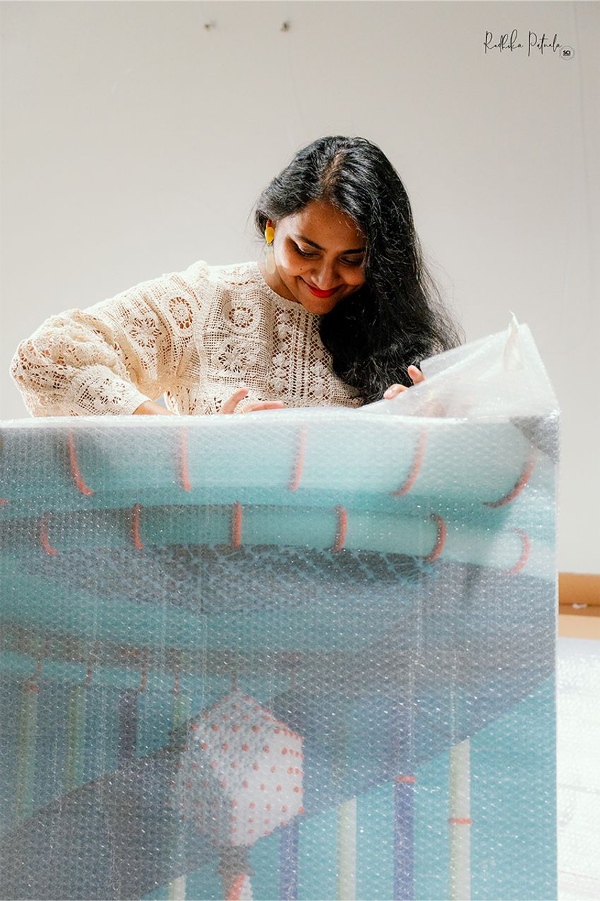 A photograph of Radhika Patnala, wearing an off-white top, with her artwork. A photograph of Radhika Patnala, wearing an off-white top, with her artwork.