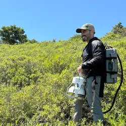 Photograph of Avi Flamholz standing in a field wearing a long black shirt and a large pack on his shoulders attached to a device in his hands used for collecting environmental samples. 