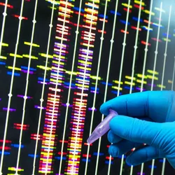 A blue-gloved hand holds a DNA sample tube in front of a computer screen that shows massively parallel sequencing data.