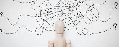 Cartoon of a wooden human-shaped figure against a white wall with several overlapping and tangled dotted lines above their head. The dashed lines end in question marks at the edge of the image. 