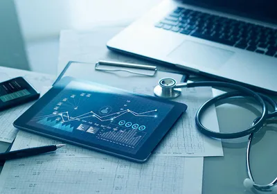 A tablet displaying patient data graphs alongside printed medical charts, a stethoscope, and a laptop.