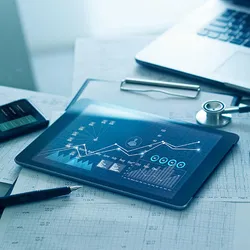 A tablet displaying patient data graphs alongside printed medical charts, a stethoscope, and a laptop.