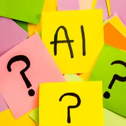 Photograph of brightly colored sticky notes arranged haphazardly. Three notes on top have question marks written in black marker on them, and a fourth sitting above these three has “AI” written on it. 