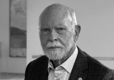 Greyscale photograph of J Craig Venter. Venter has a short white beard and  is wearing a patterned suit jacket with a gold pin on one lapel over a light blue button-down shirt. 