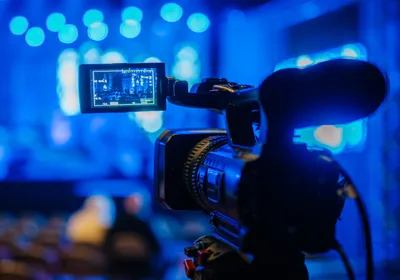 Video camera on tripod is focused on live performance stage illuminated by blue lights, highlighting science education through documentaries and films.