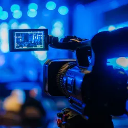 Video camera on tripod is focused on live performance stage illuminated by blue lights, highlighting science education through documentaries and films.