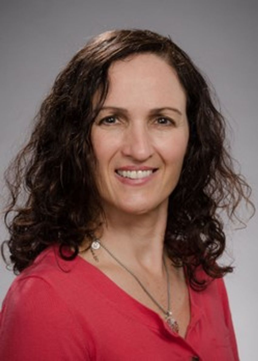 Photograph of Deborah Fuller, an immunologist at the University of Washington. Fuller is sitting in front of a grey background and is wearing a pink top. She has curly dark hair and is smiling at the camera.