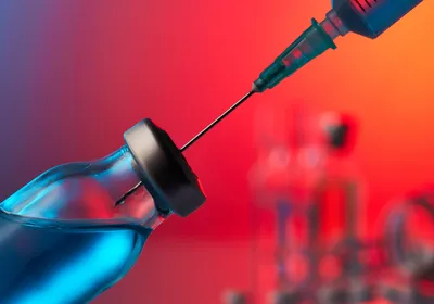 Image of a syringe inserted into a vial with a bluish liquid against a red background. 