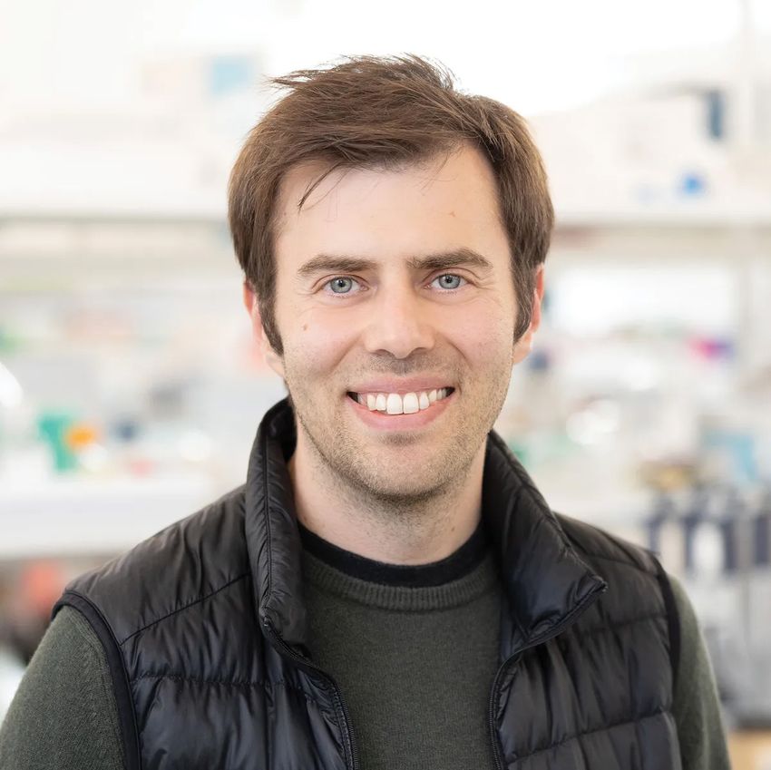Photograph of Daniel Lin-Arlow in front of a blurred background of a research lab. Lin-Arlow has short light brain hair and is wearing a grey long-sleeve shirt with a puffy black vest over it.