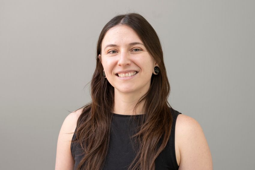 Jessie Davis smiles, wearing a black sleeveless top against a grey backdrop. She has long brown hair.