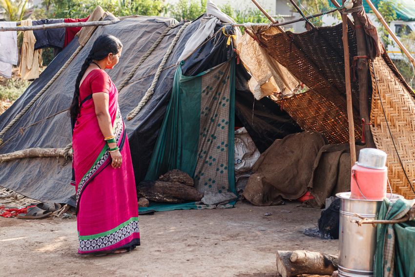 A community healthcare worker stands outside a heat-exposed shelter made up of blankets and cloths held up on posts. A community healthcare worker stands outside a heat-exposed shelter made up of blankets and cloths held up on posts.
