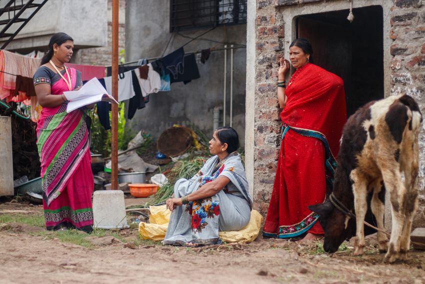 A community healthcare worker holds an informational booklet while talking to a women sitting down and another woman standing in a doorway. A cow stands next to them. A community healthcare worker holds an informational booklet while talking to a women sitting down and another woman standing in a doorway. A cow stands next to them.