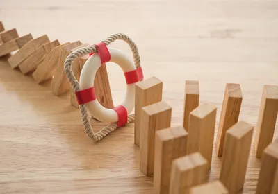 Photograph of wooden domino pieces on a table. A line of them extending from the top left corner has fallen over. A red and white inflatable ring in a rope square stops the fallen dominoes from knocking over a larger group of them on the bottom right of the image. The image represents the protective effect of preventative measures. 