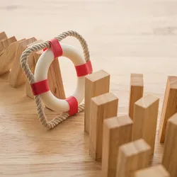 Photograph of wooden domino pieces on a table. A line of them extending from the top left corner has fallen over. A red and white inflatable ring in a rope square stops the fallen dominoes from knocking over a larger group of them on the bottom right of the image. The image represents the protective effect of preventative measures. 
