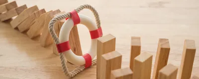 Photograph of wooden domino pieces on a table. A line of them extending from the top left corner has fallen over. A red and white inflatable ring in a rope square stops the fallen dominoes from knocking over a larger group of them on the bottom right of the image. The image represents the protective effect of preventative measures. 