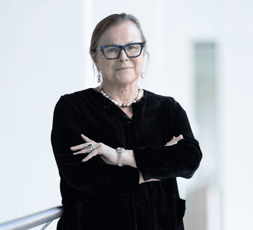 Fiona Wood is smiling with her arms folded across her chest, wearing a black jacket and glasses.