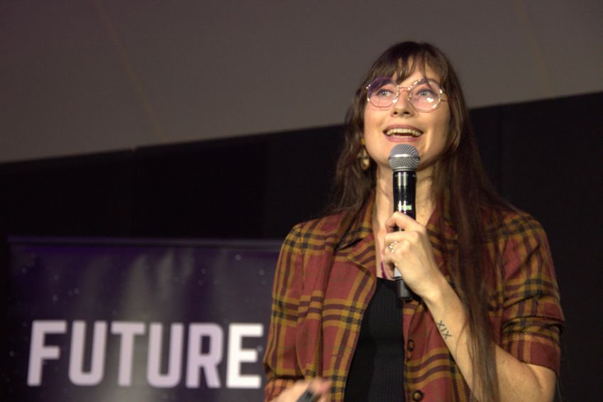 Alise Fox is wearing a red plaid jacket and glasses, holding a microphone on a stage as she talks about math.
