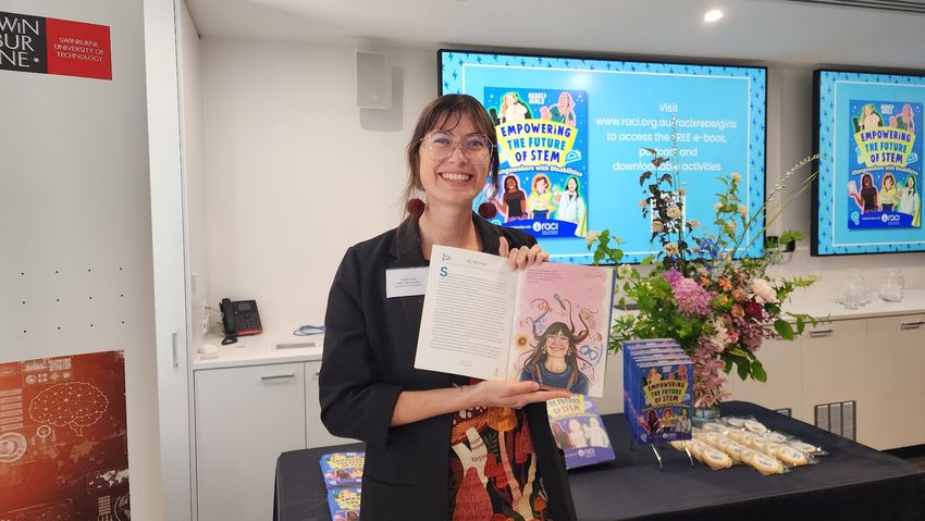 Alise Fox holds a book with her own picture in it, standing in front of a presentation slide with the words ‘Empowering the Future of STEM’.