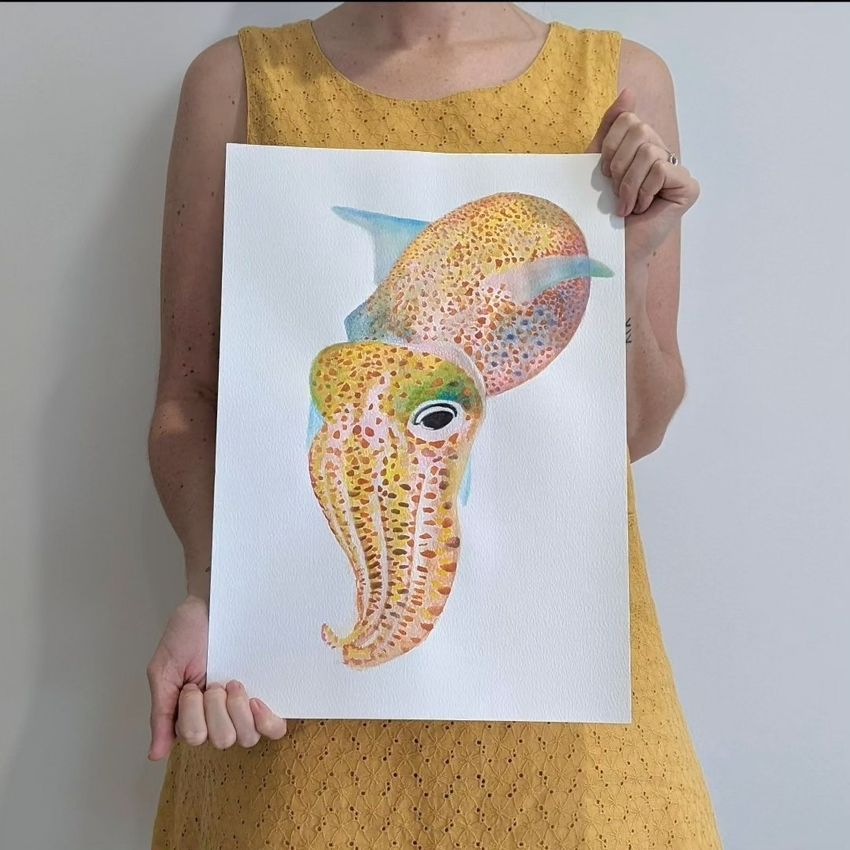 Alise Fox is wearing a yellow dress and is holding a watercolor painting of a colorful bobtail squid.