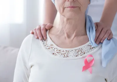 Breast cancer awareness concept with pink ribbon on a sick, older woman and supportive hands on her shoulder.
