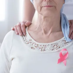 Breast cancer awareness concept with pink ribbon on a sick, older woman and supportive hands on her shoulder.
