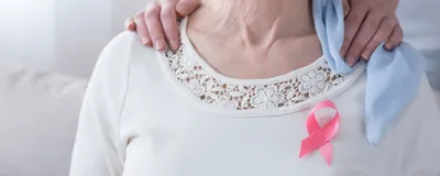 Breast cancer awareness concept with pink ribbon on a sick, older woman and supportive hands on her shoulder.