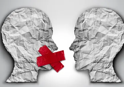 3D illustration of two human head shapes made on crumpled paper facing each other on a grey background. The head on the left has red tape covering its mouth, representing censorship.