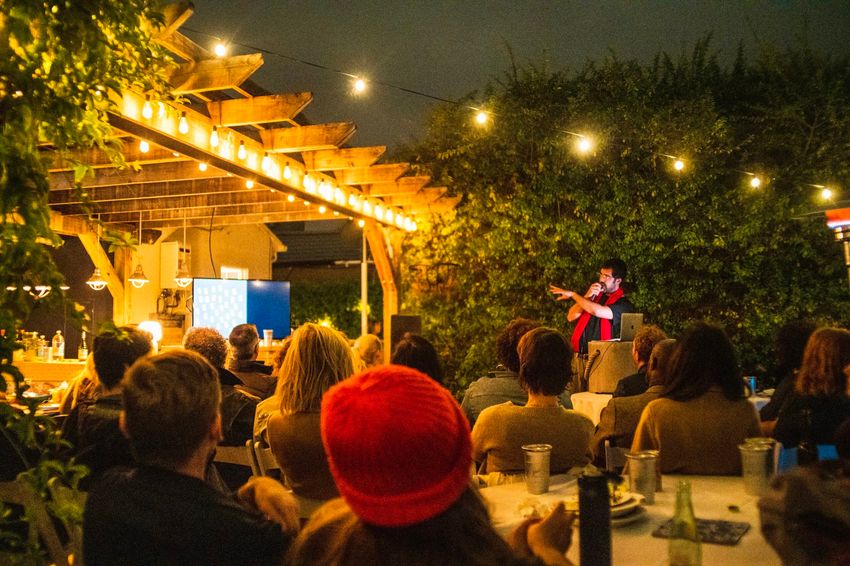 Photograph of a man standing in front of a seated audience outdoors at night indicating to a video screen and holding a microphone.