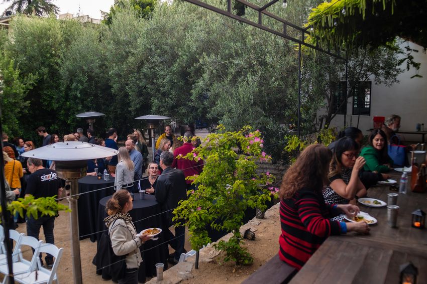 Photograph of an outdoor event with a raised section with a picnic table in the foreground and more cocktail tables arranged on the ground in the rest of the photo. People are gathered at the tables with plates of food and are talking.