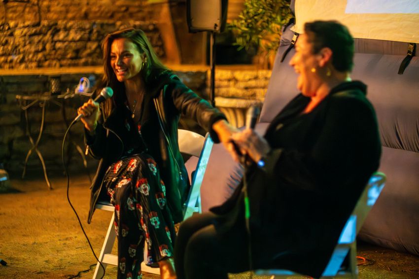 Photograph of two women sitting in folding chairs and holding microphones, laughing together.