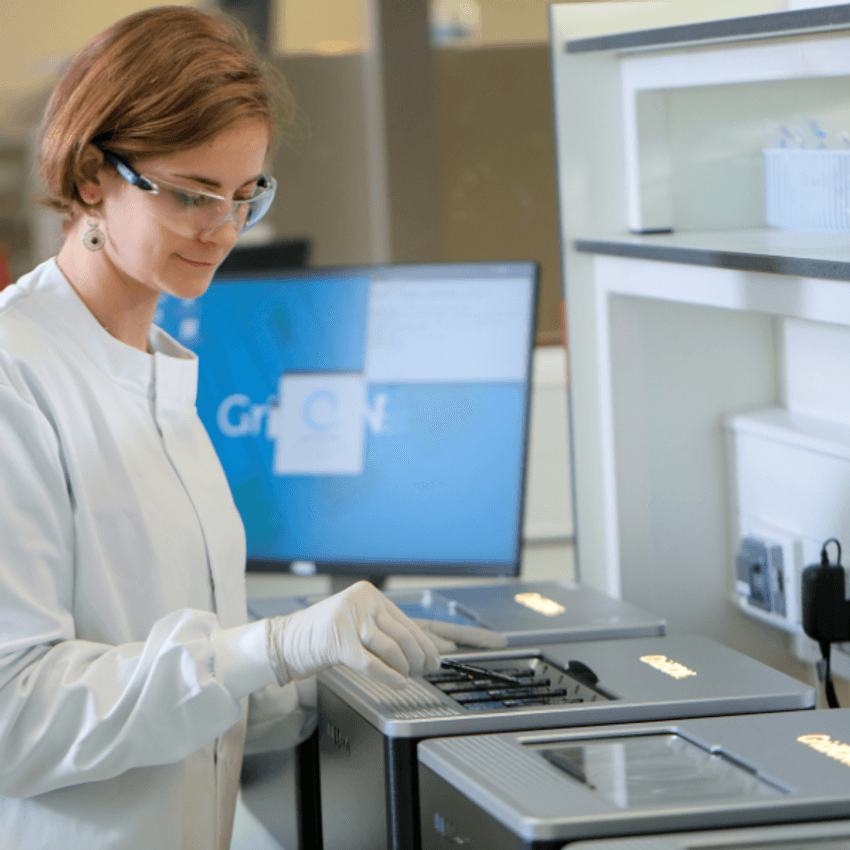 A scientist in a white lab coat loading a MinION™ flow cell into a GridION™ sequencer. A computer monitor in the background shows a GridION homepage. A scientist in a white lab coat loading a MinION™ flow cell into a GridION™ sequencer. A computer monitor in the background shows a GridION homepage.