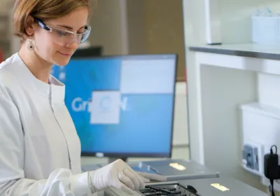 A scientist in a white lab coat loading a MinION™ flow cell into a GridION™ sequencer. A computer monitor in the background shows a GridION homepage.