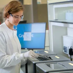 A scientist in a white lab coat loading a MinION™ flow cell into a GridION™ sequencer. A computer monitor in the background shows a GridION homepage.