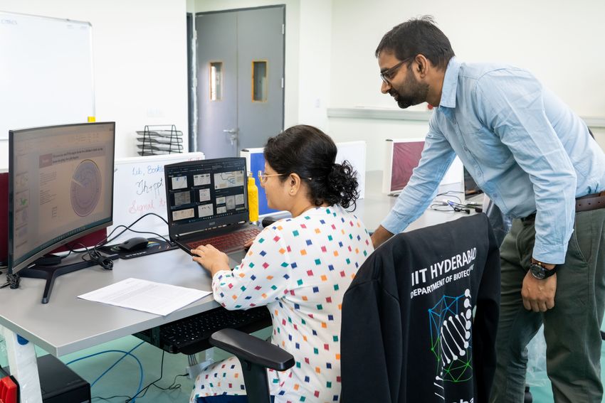 A photograph of Utkarsha Mahanta sitting in front of a computer, on a chair with a jacket that says IIT Hyderabad as Gaurav Sharma stands behind her.