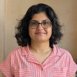 Sheetal Potdar wears a pink shirt and glasses in front of a brown wall.