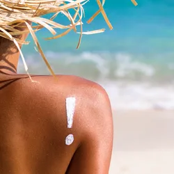 An exclamation mark is drawn on a woman’s bare shoulder in sunscreen as she sits on a beach wearing a straw hat.