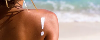 An exclamation mark is drawn on a woman’s bare shoulder in sunscreen as she sits on a beach wearing a straw hat.