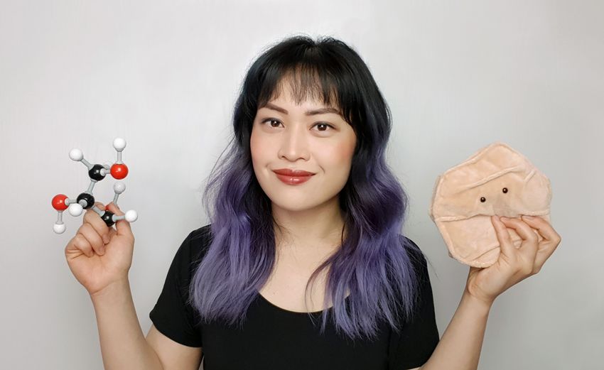 Michelle Wong smiles, holding a plushie of a human skin cell in one hand and a three-dimensional model of a glycerin molecule in another. Her hair is black with purple ombre ends. Michelle Wong smiles, holding a plushie of a human skin cell in one hand and a three-dimensional model of a glycerin molecule in another. Her hair is black with purple ombre ends.