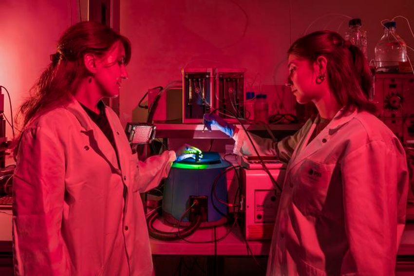 Two researchers wear lab coats while bathed in red light with a scientific instrument with green light emanating from it in the center. Two researchers wear lab coats while bathed in red light with a scientific instrument with green light emanating from it in the center.